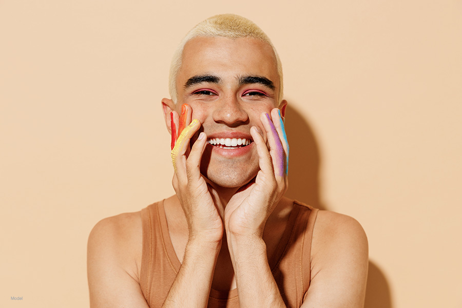 Young smiling happy fun blond latin gay man 20s wearing beige tank shirt posing with make up fingers painted in rainbow flag colors hold face isolated on plain light ocher background studio portrait. a person with rainbow's painted on their fingers